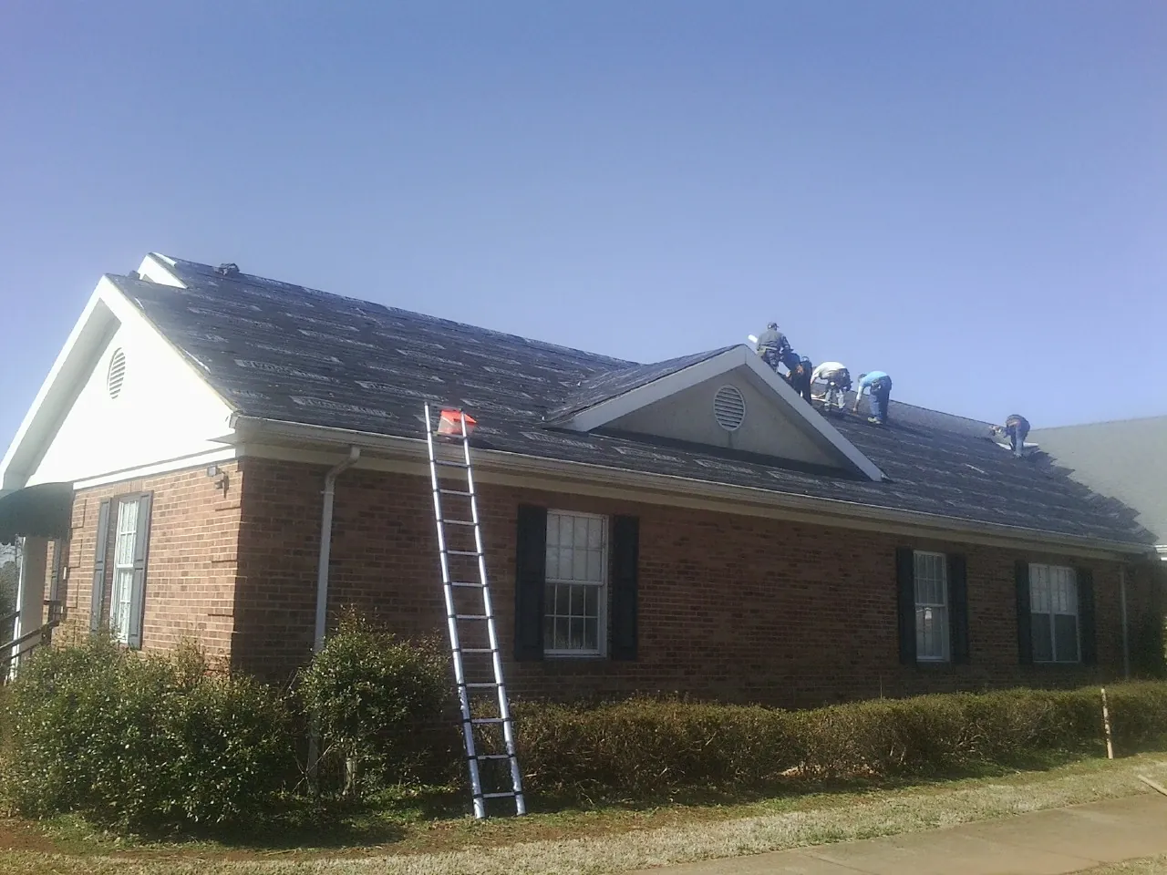 Crew tearing shingles off a brick ranch roof with a ladder set — Metal Roof Repair work by our crew in Otley