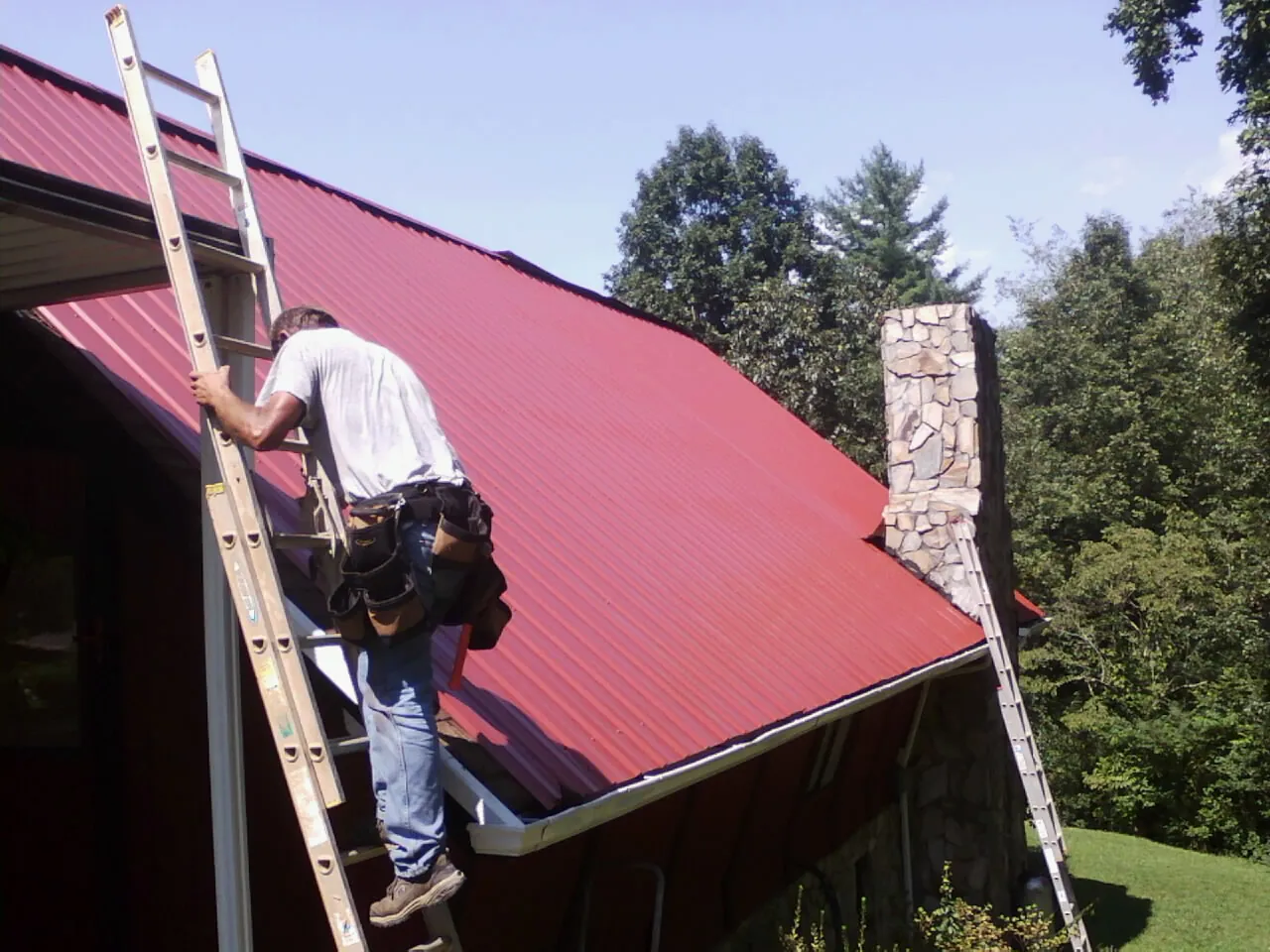 Roofer on a ladder beside a red metal roof and stone chimney — Soffit & Fascia Repair by Roofing Brothers in Otley
